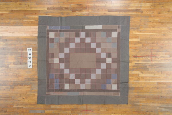 Muted gray and taupe patchwork rug with red stitched seams lies flat on distressed hardwood flooring.