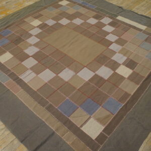 Patchwork textile in earthy neutral colors features visible contrast stitching and rests on distressed wooden floorboards.