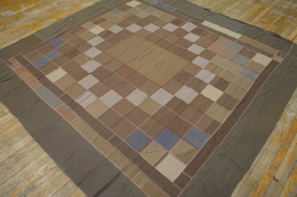 Patchwork textile in earthy neutral colors features visible contrast stitching and rests on distressed wooden floorboards.