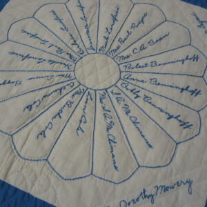 Low-pile cream textile featuring blue embroidered names within a circular petal design and quilted diamonds.