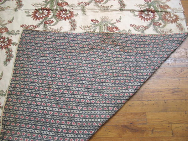 Cream botanical textile folded to show gray and red geometric backing resting on distressed wood flooring.