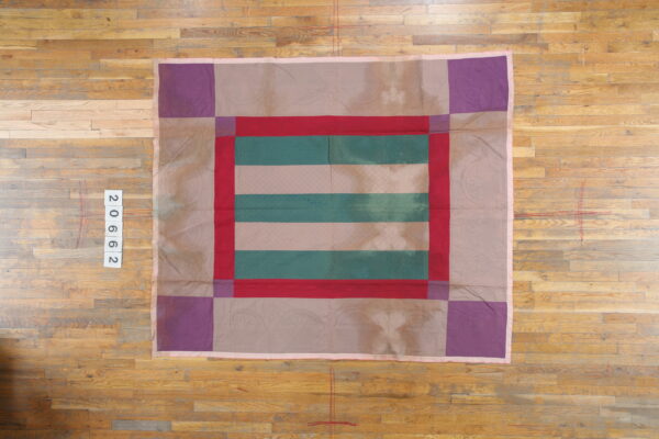 Square textile with red, teal, and mauve striped center, purple corners, and low-contrast patterned borders on a wood floor.