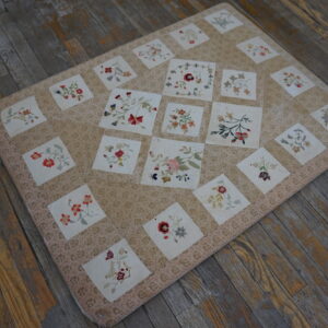 Beige floral textile with squares of multicolored embroidered blooms covers wide, distressed gray wooden boards.
