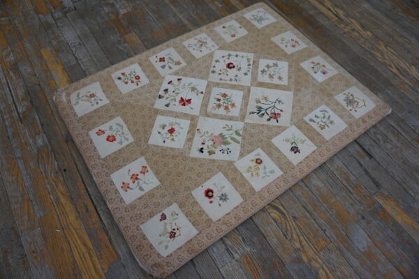 Beige floral textile with squares of multicolored embroidered blooms covers wide, distressed gray wooden boards.