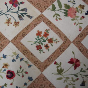 Close-up of a diamond patchwork textile pattern featuring embroidered pink, red, orange, and blue florals on a cream ground.