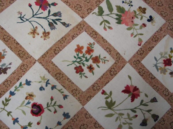 Close-up of a diamond patchwork textile pattern featuring embroidered pink, red, orange, and blue florals on a cream ground.