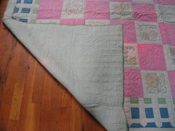 Quilted textile with pink and embroidered white floral blocks rests partially on vertical hardwood planks.