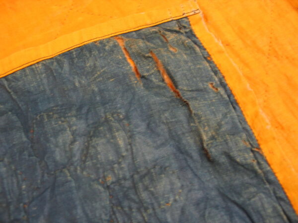 Distressed blue textile with visible tears meets a plain golden-orange fabric border with black stitching.