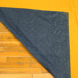 A bright yellow rug corner is folded, revealing a quilted dark blue backing on hardwood flooring.