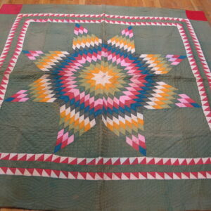 A textured, sage green textile rug lies on a wood floor featuring a bright, multicolor starburst medallion and sawtooth border.