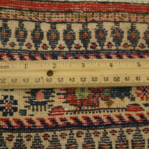 Detailed view of a beige, red, and blue patterned rug with a wooden ruler providing scale reference.