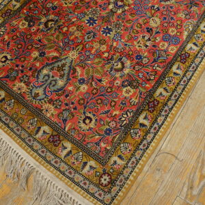 Intricate floral rug featuring a coral-red field and golden border on worn, light wood planks.