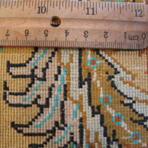 Detailed view of low-pile woven rug in gold, black, teal, and white with a wooden ruler for scale.