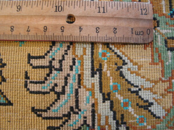 Detailed view of low-pile woven rug in gold, black, teal, and white with a wooden ruler for scale.