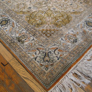 The corner of a low-pile ivory rug with ornate gold and blue floral patterns on distressed hardwood.