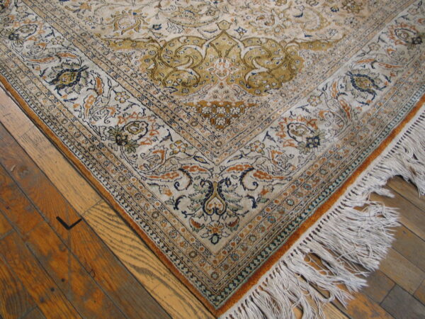 The corner of a low-pile ivory rug with ornate gold and blue floral patterns on distressed hardwood.
