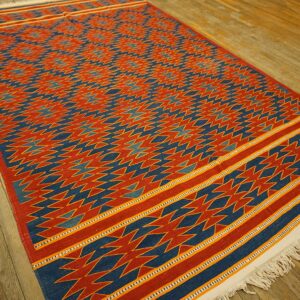 Red and blue flatweave rug with a detailed geometric pattern and striped borders rests on wood floorboards.