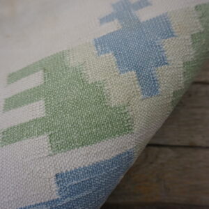 Curled rug edge displays a textured geometric pattern in cream, light blue, and sage green on wood.