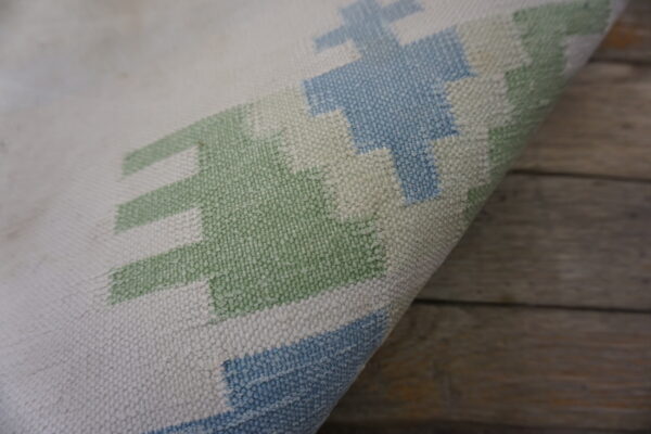 Curled rug edge displays a textured geometric pattern in cream, light blue, and sage green on wood.