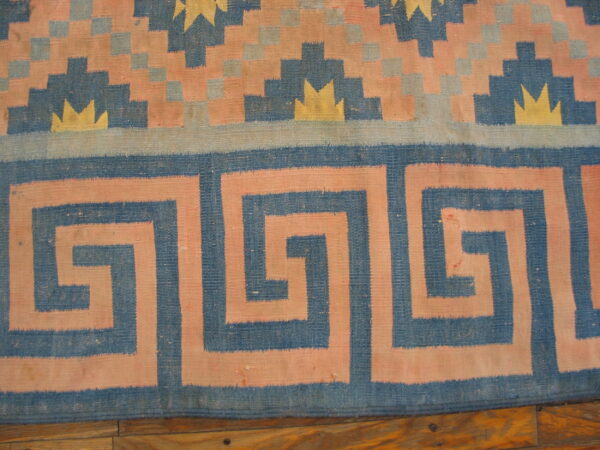 Geometric flatweave rug in blue, pink, and yellow with a meander border over wood flooring.