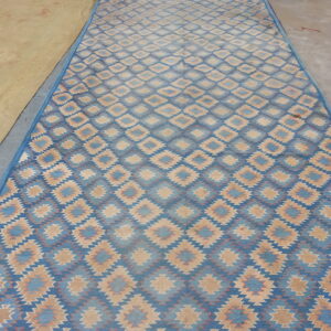 Blue and tan flatweave rug with repeating serrated diamond patterns on a concrete floor.