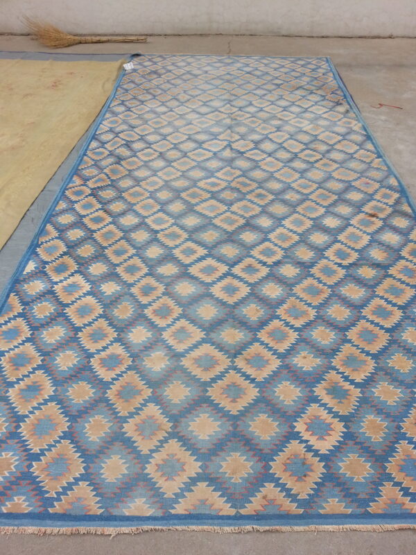 Blue and tan flatweave rug with repeating serrated diamond patterns on a concrete floor.