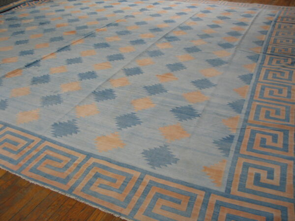 Blue and peach flatweave rug with repeating jagged diamond motifs and a wide meander border on a wood floor.