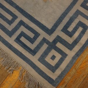 A flatweave rug with a blue and beige Greek Key pattern and fringe rests on distressed wood flooring.