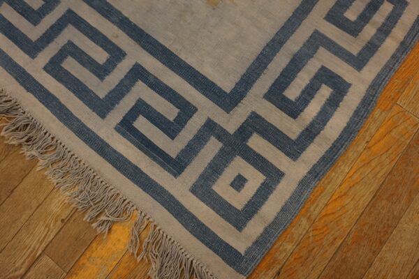 A flatweave rug with a blue and beige greek key pattern and fringe rests on distressed wood flooring.
