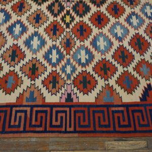 Flatweave rug featuring serrated rust, navy, and blue geometric diamonds and a contrasting Greek key border on wood floor.