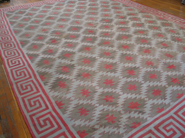 A large, low-pile rug featuring red, gray, and white sawtooth patterns and a greek key border on wood flooring.