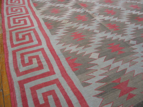 Low-pile geometric rug in pink, gray, and light blue with a greek key border on wood flooring.