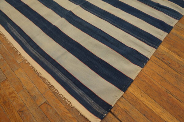 A flatwoven rug with wide cream and indigo stripes and fringe rests on rustic wood plank flooring.