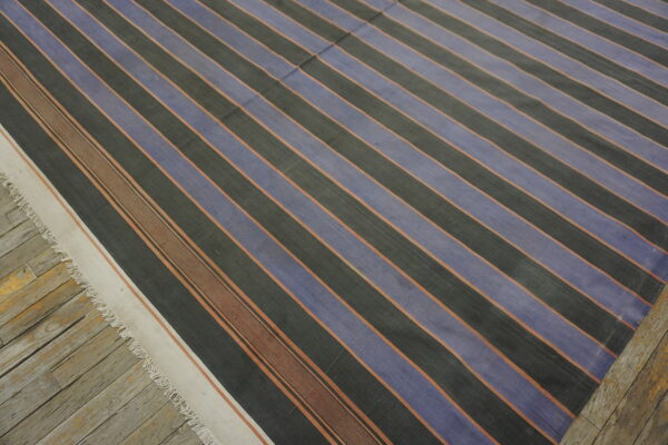 Striped flatweave rug featuring blue and dark grey bands accented by thin rust lines on weathered wood flooring.