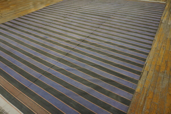 Wide blue and black striped flatweave rug with thin separating lines rests on a rustic wood floor.