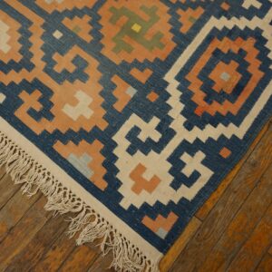 Geometric blue, cream, and orange flatweave rug with fringe resting on worn plank hardwood flooring.
