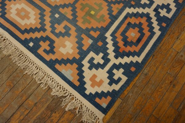 Geometric blue, cream, and orange flatweave rug with fringe resting on worn plank hardwood flooring.