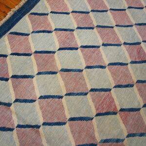 Flatweave rug with an optical cube pattern in muted red, blue, and light gray on wood flooring.