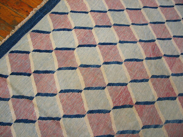 Flatweave rug with an optical cube pattern in muted red, blue, and light gray on wood flooring.
