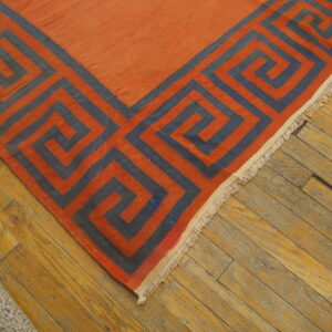 Orange flatweave rug featuring a dark blue Greek Key border rests on distressed golden wood floorboards.