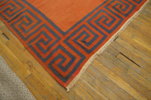 Orange flatweave rug featuring a dark blue greek key border rests on distressed golden wood floorboards.