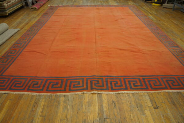 Flat-weave terracotta rug featuring a wide slate-blue geometric key border on worn wood plank flooring.