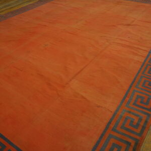 A reddish-orange flatweave rug featuring a gray Greek key border rests on worn hardwood flooring.