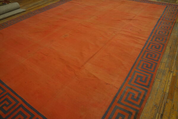 A reddish-orange flatweave rug featuring a gray greek key border rests on worn hardwood flooring.