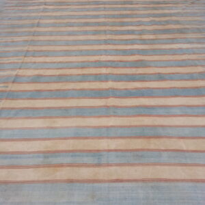 Large flatweave rug with horizontal stripes in pale blue, cream, and rust, lying on a concrete floor.