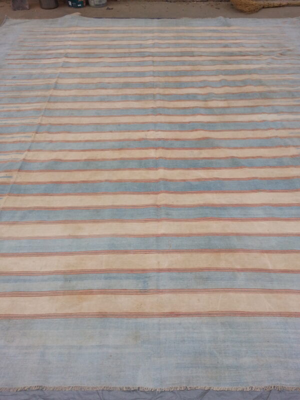 Large flatweave rug with horizontal stripes in pale blue, cream, and rust, lying on a concrete floor.