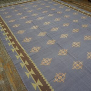 Blue-gray flatweave rug with yellow geometric patterns and brown borders rests on distressed wood flooring.