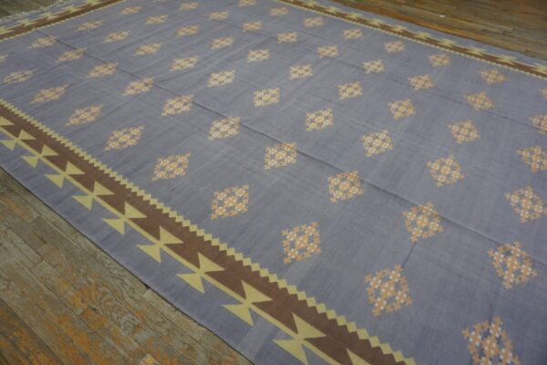Flat-woven rug with gray-blue field, checkered diamonds, and a brown geometric border on wood floors.