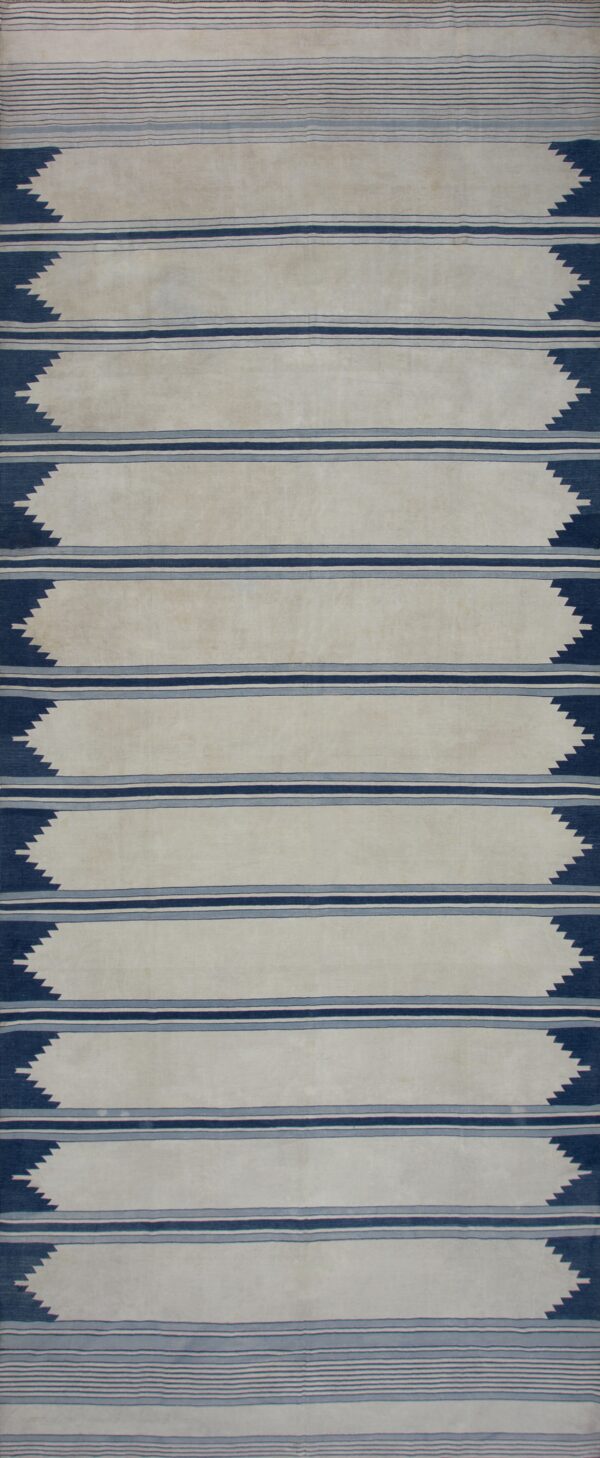 Off-white flatweave rug with vertical triple blue stripes and dark blue serrated zigzags on wood flooring.