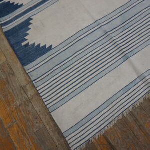 White flatweave rug with blue stripe and sawtooth patterns rests on distressed wooden floorboards.
